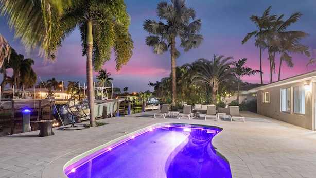 A swimming pool with a unique shape, surrounded by palm trees and lounge chairs, with a view of a building in the background.