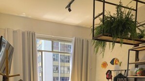 A compact living space with a bed, a dining table, and a small kitchen area. There is a window with curtains, a fan, and a shelf with decorative items. - Sua Casa no Rio-Lapa-Lindo Studio (Rio de Janeiro)