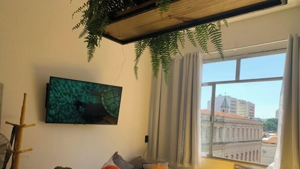 A room with a bed, a desk, a chair, a window with a view of buildings, and a plant hanging from the ceiling. - Sua Casa no Rio-Lapa-Lindo Studio (Rio de Janeiro)
