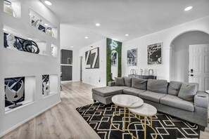 Un soggiorno moderno con un divano angolare grigio, un tavolino rotondo e un tappeto decorativo. Si vedono mensole con oggetti d'arredo e opere d'arte incorniciate alle pareti.