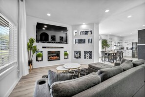 Un salon moderne avec un canapé gris, une cheminée et un coin repas, la cuisine étant visible au fond.