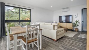 A modern living room with a wooden dining table, white chairs, a beige sofa, a flat-screen TV, and a large window offering a view of greenery. - Villa Piedade by Homie (Ponta do Sol)