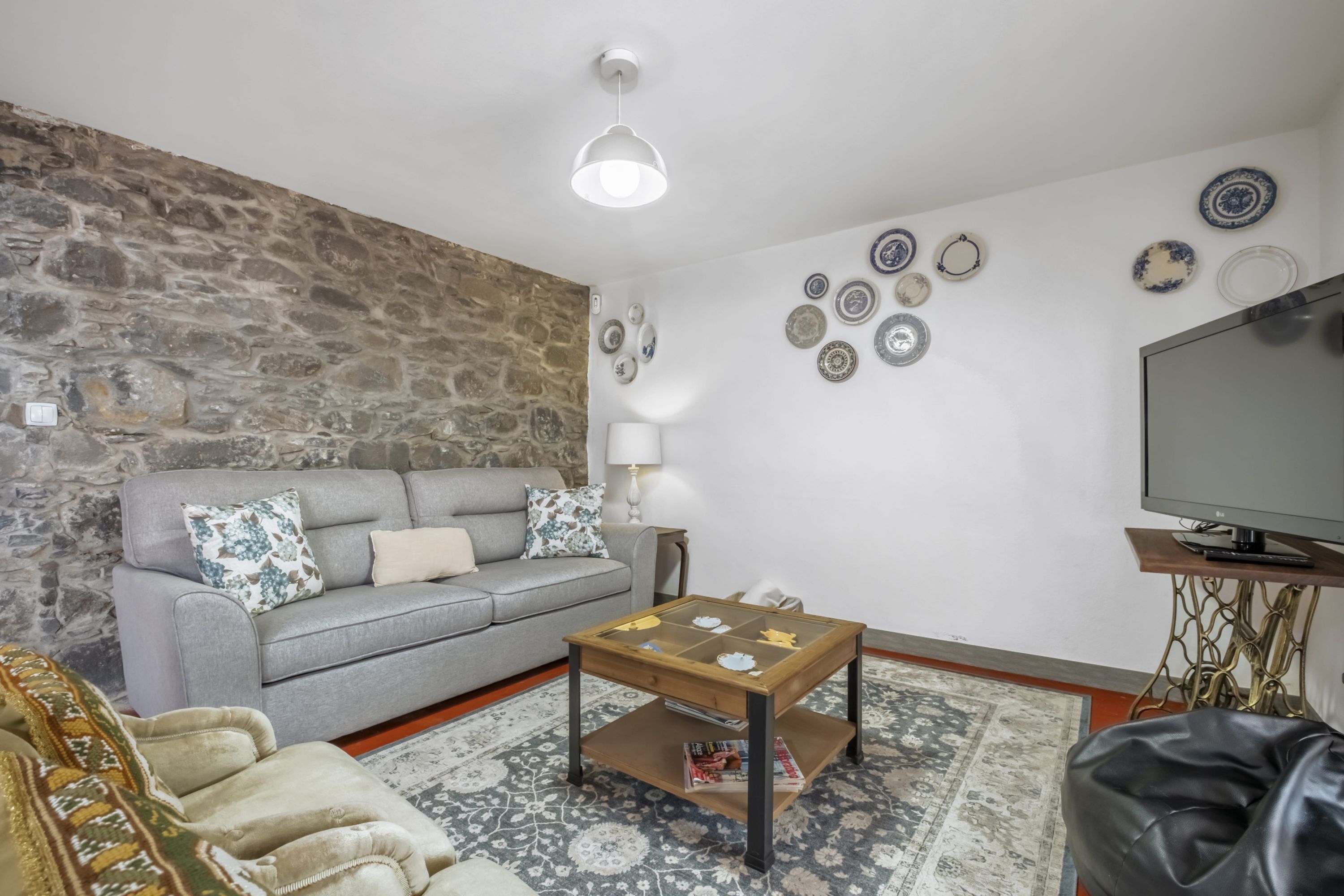 A living room with a stone wall, grey sofa, patterned rug, coffee table, and a television on a wooden stand.