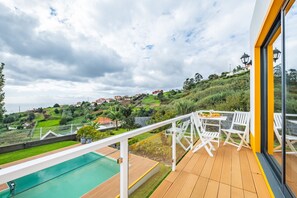 Un balcon avec une piscine, des chaises blanches et une vue sur une colline parsemée de maisons.