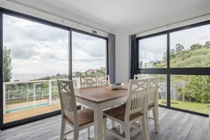 Un espace repas avec une table en bois et des chaises blanches, de grandes fenêtres offrant une vue sur un paysage verdoyant et des bâtiments, ainsi qu’un balcon doté d’une rambarde en verre.