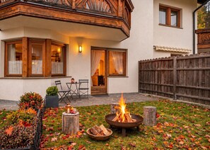 Un rincón al aire libre acogedor con una chimenea, terraza de madera y vistas a un edificio con amplias ventanas y un balcón.