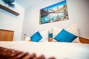 Un dormitorio con cama, almohadas, mesita de noche, lámpara y un cuadro de un paisaje montañoso.
