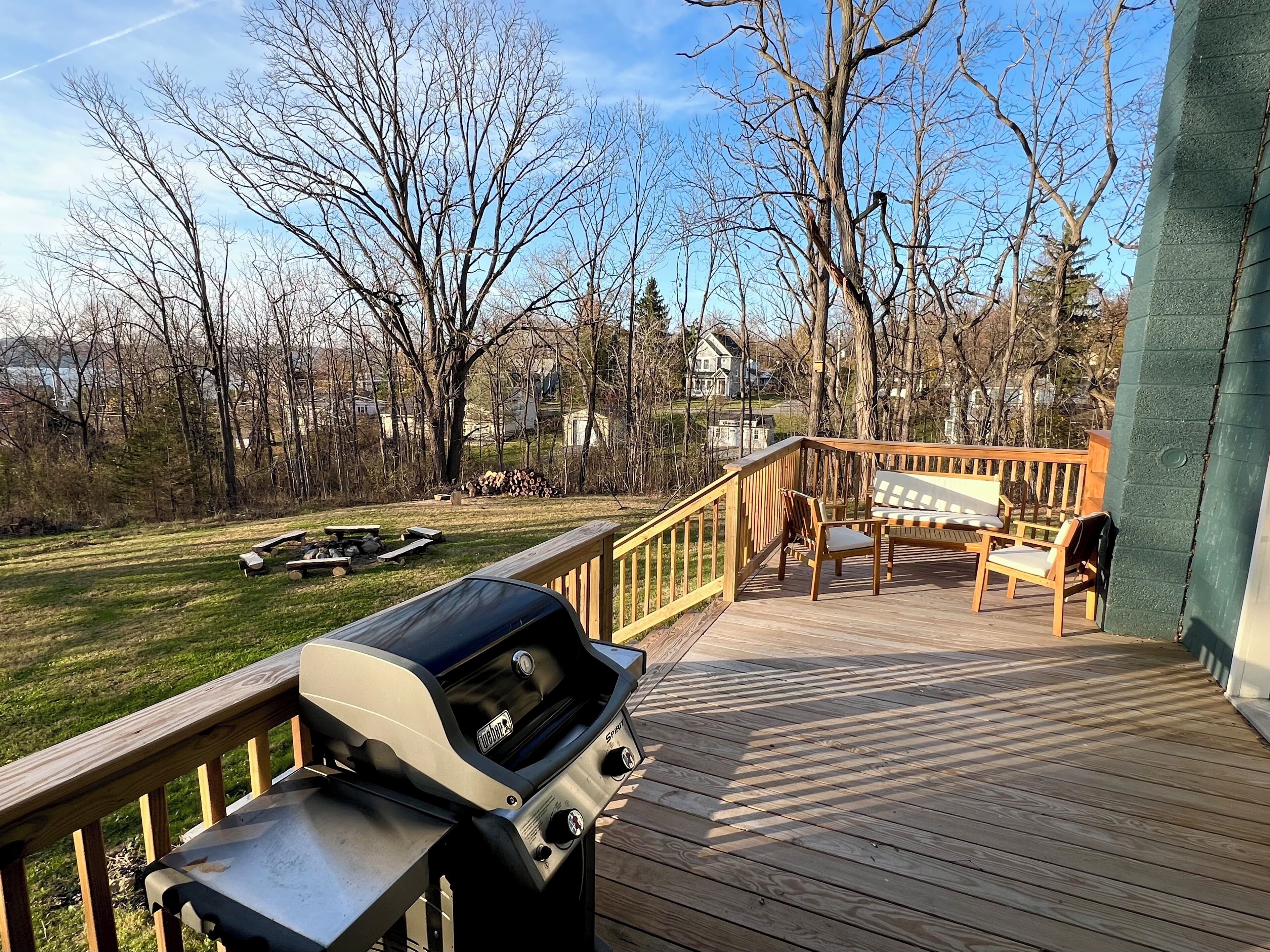 A wooden deck with a grill, outdoor furniture, and a view of a yard with trees and houses in the background.