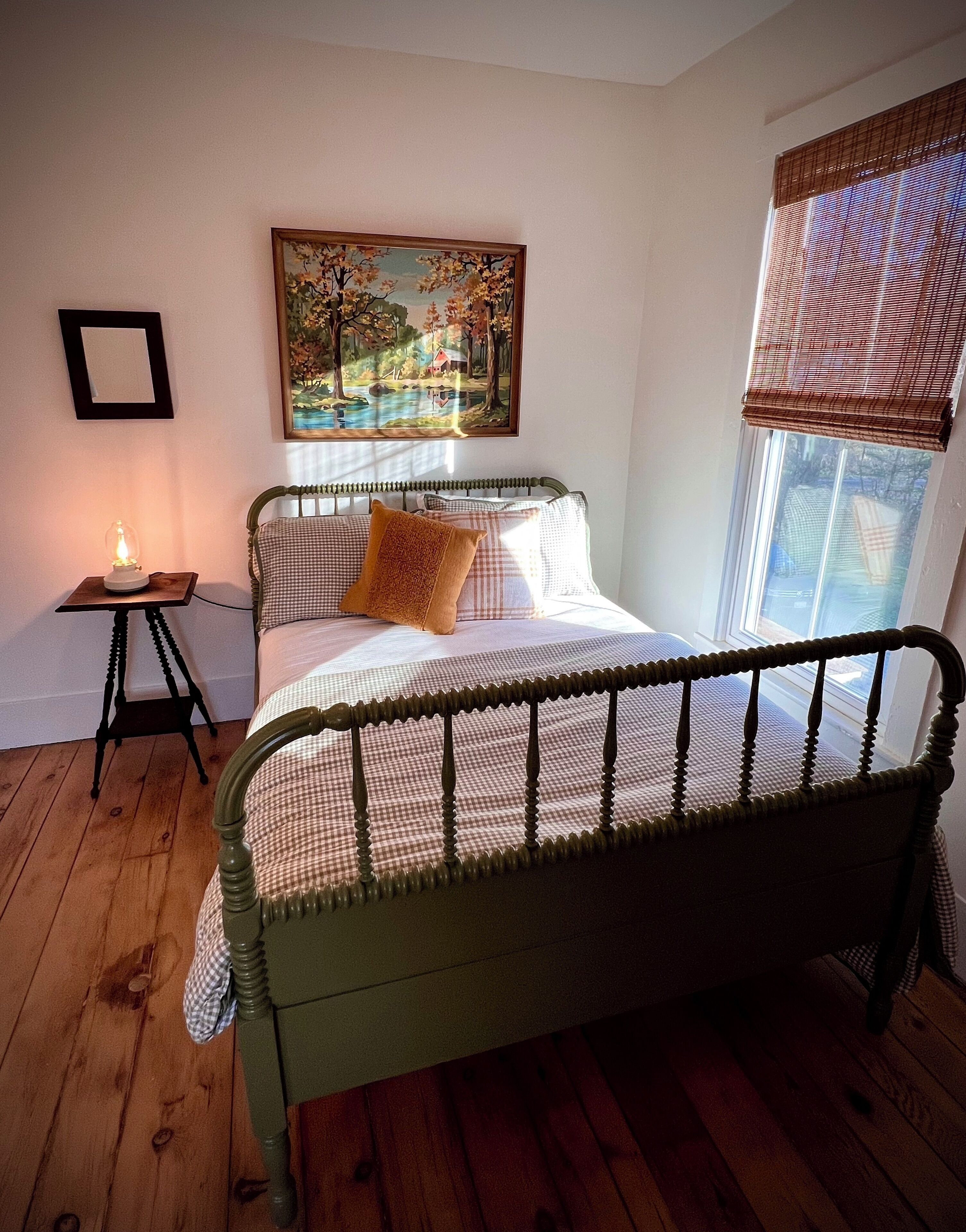 Une chambre avec un plancher en bois, un lit avec un cadre en métal, une table de chevet avec une lampe, un tableau au mur et une fenêtre avec des stores.