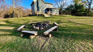 Une maison en pierre à deux étages, dotée d’une terrasse en bois et d’un foyer extérieur dans le jardin.