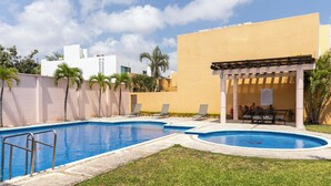 Ein Swimmingpool mit Liegestühlen und einer Pergola, umgeben von Palmen und einem Gebäude mit gelber Fassade.
