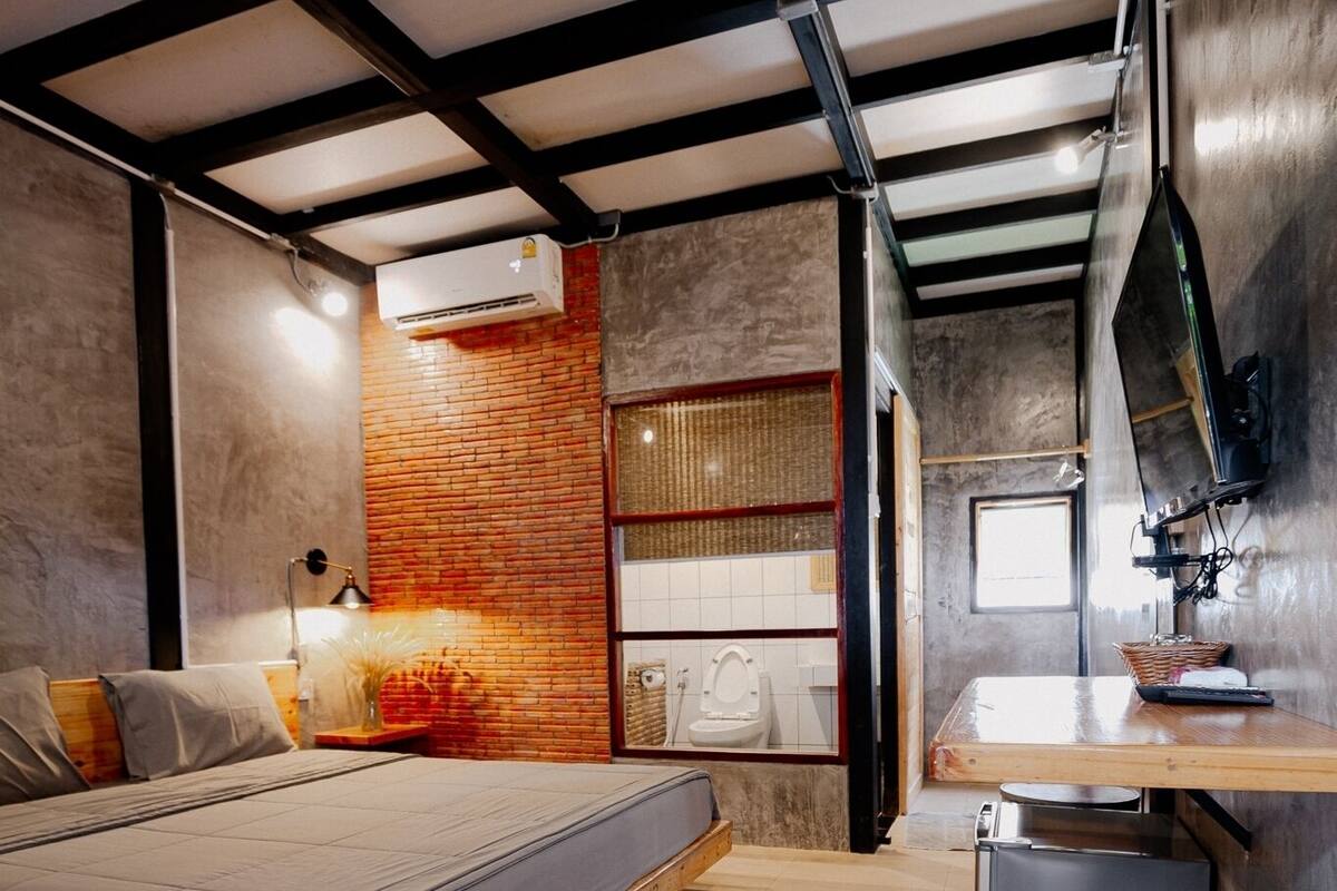 A compact bedroom with a bed, a small bedside table, a nightstand, a wall-mounted shelf, and a window.