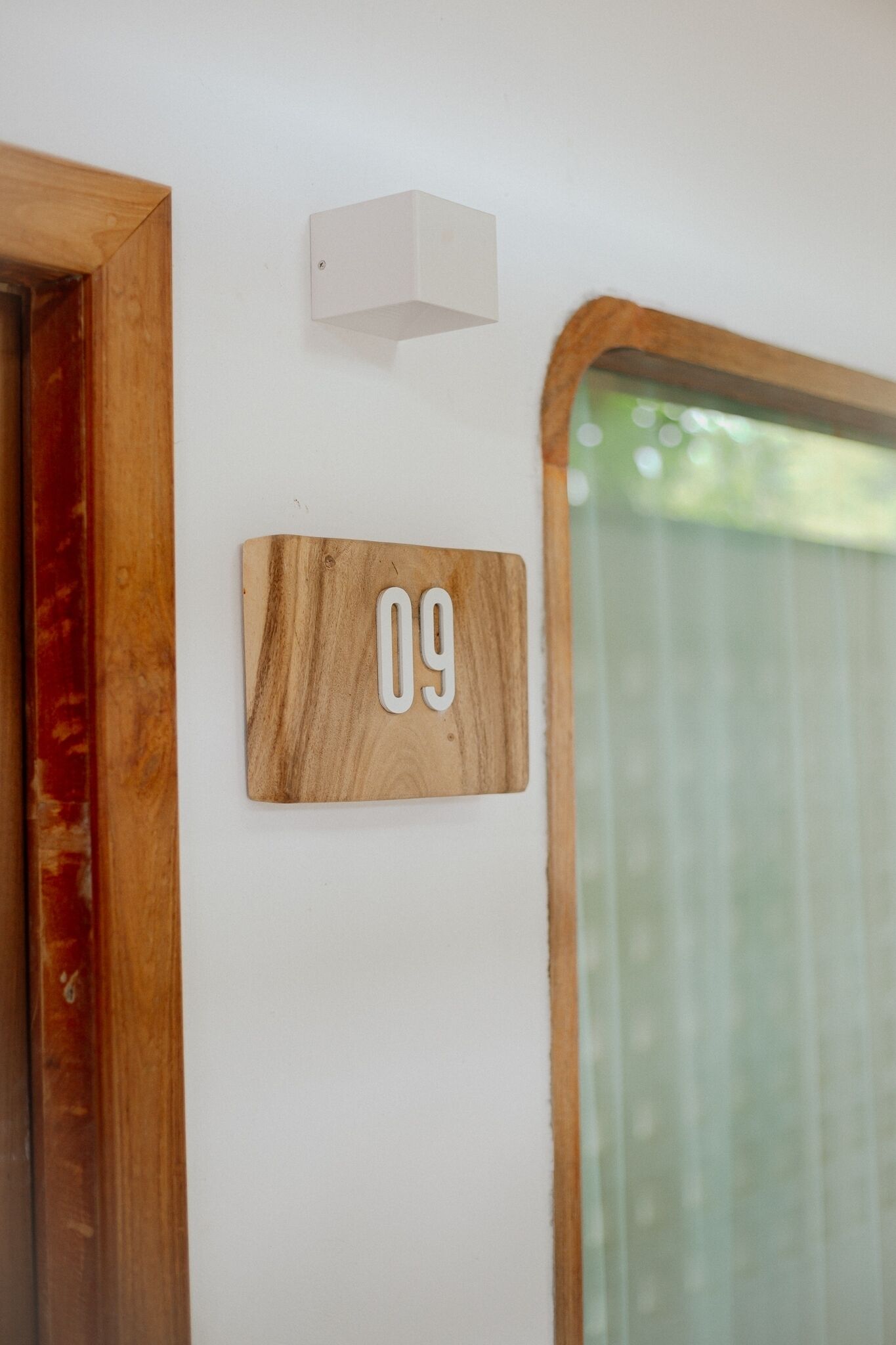 Una placa de madera con el número 09 en una pared blanca, una pequeña luminaria blanca encima y una ventana con marco de madera a la derecha.