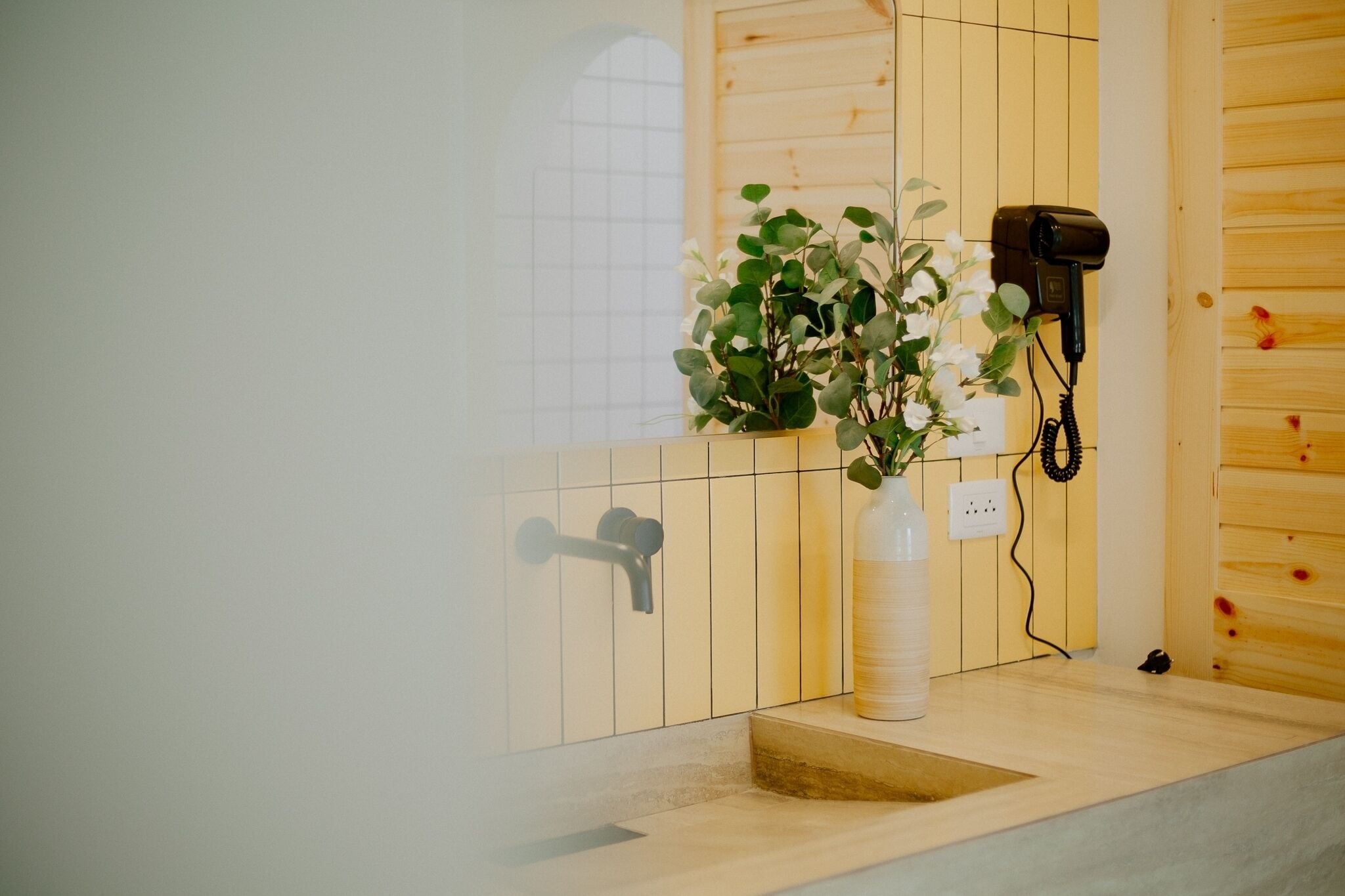 A bathroom with a wooden wall, a white tiled wall, a sink, a hairdryer, and a potted plant.
