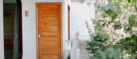 A wooden door with a glass window to the left, a white wall, and a white railing with greenery in the foreground.