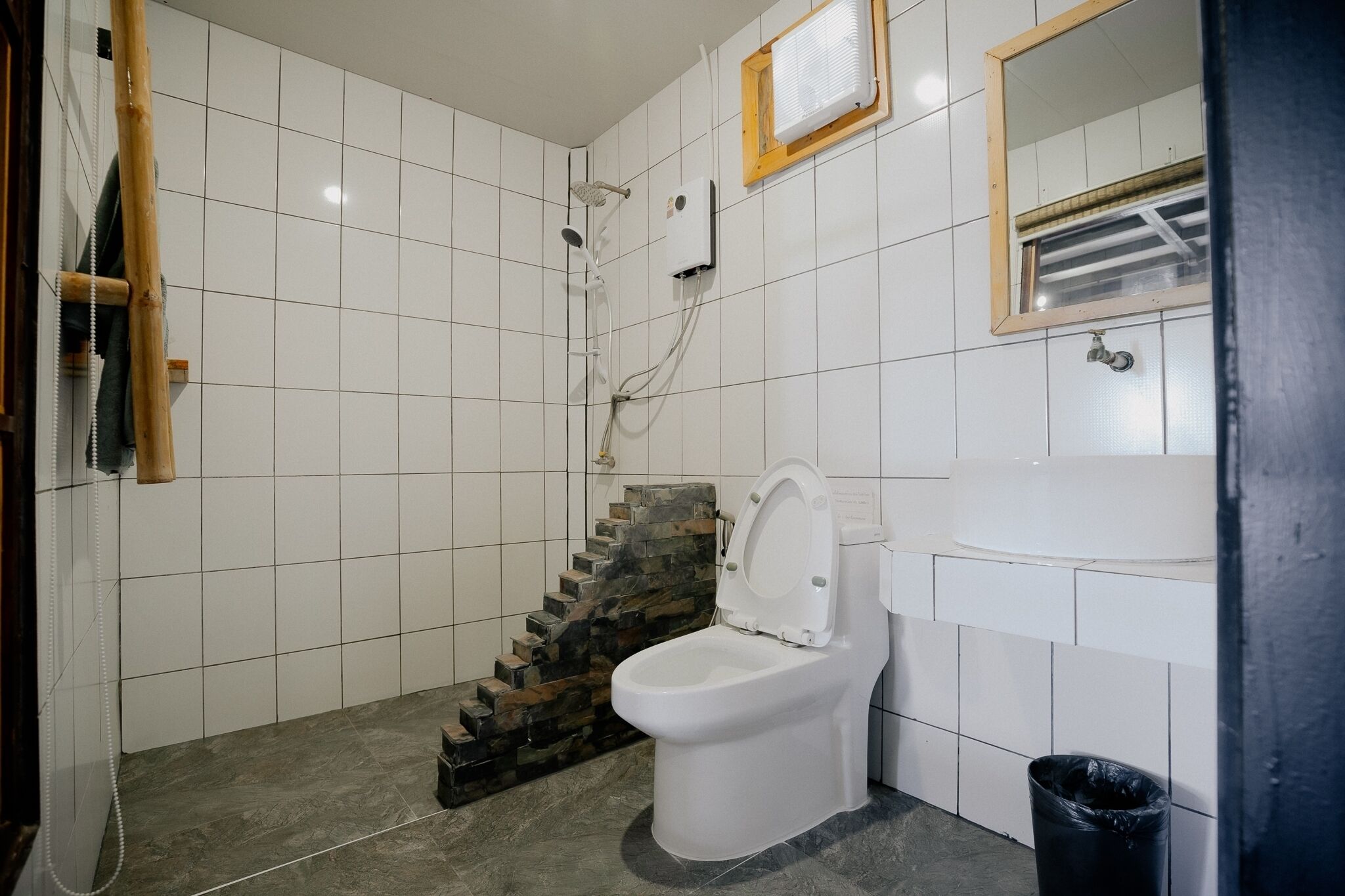 A bathroom with white tiled walls, a white toilet, a sink, a mirror, and a shower with a stone-tiled wall.