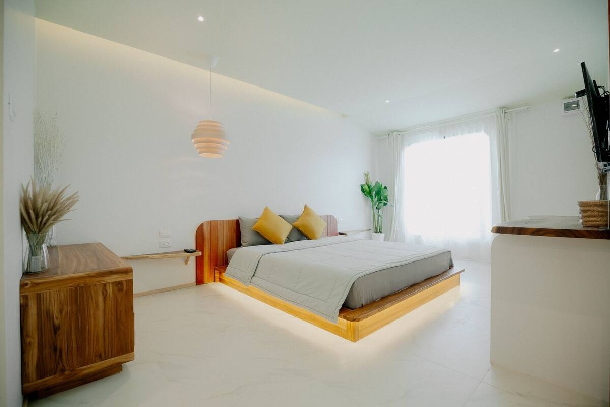 A modern bedroom with a wooden bed frame, a bedside table, a hanging light fixture, and a window with curtains.