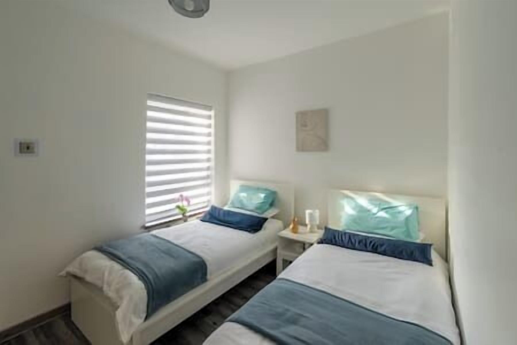 Ein kleines, sauberes Zimmer mit zwei Einzelbetten, einem Nachttisch mit Lampe und einem Fenster mit Jalousien.