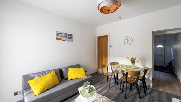 A modern living room with a grey sofa, yellow cushions, a dining table with chairs, a small white side table, and a hanging light fixture.