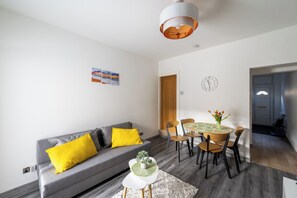 A modern living room with a grey sofa, yellow cushions, a dining table with chairs, a small white side table, and a hanging light fixture.