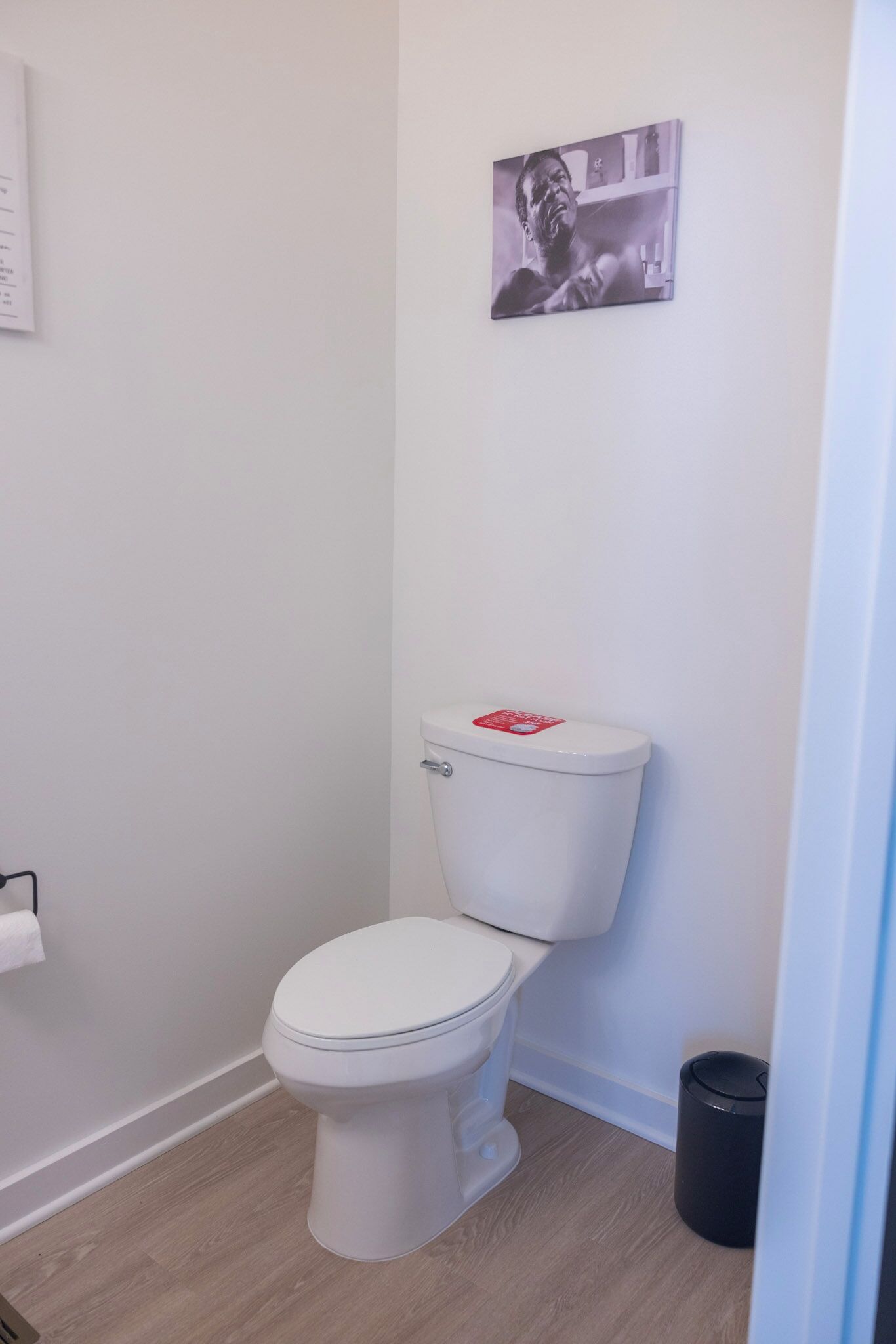 A small, clean bathroom with a white toilet, a black trash bin, and a framed picture on the wall.