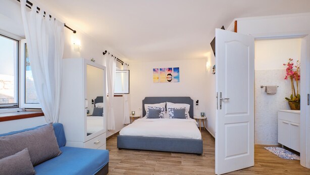 Um quarto com cama, mesa de cabeceira, penteadeira, espelho, janela com cortinas e porta para outro cômodo.