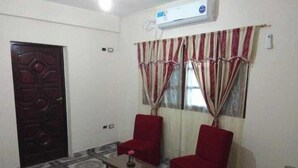 Una habitación con alfombra estampada, dos sillas rojas, una mesita con un jarrón, una puerta de madera oscura y una ventana con cortinas a rayas.