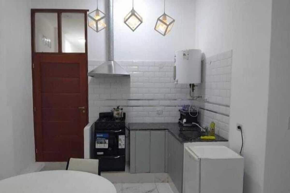 Una cocina compacta con salpicadero blanco de azulejos, una estufa, un fregadero y un refrigerador. Hay luces colgantes sobre la encimera.