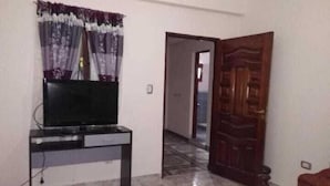 Una habitación con una televisión sobre un mueble bajo, una alfombra estampada, una puerta de madera y un pasillo que conduce a otra habitación.