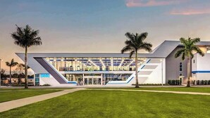A modern building with a glass facade, palm trees, and a blue logo.
