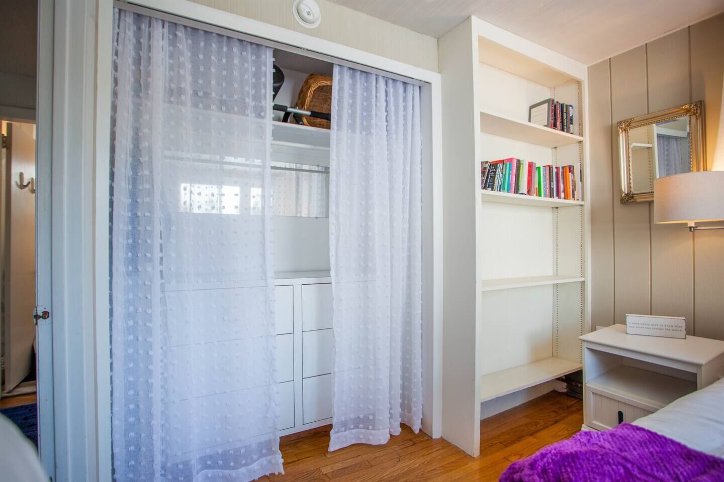 A bedroom with a wardrobe, a bedside table, a bookshelf, and a mirror.
