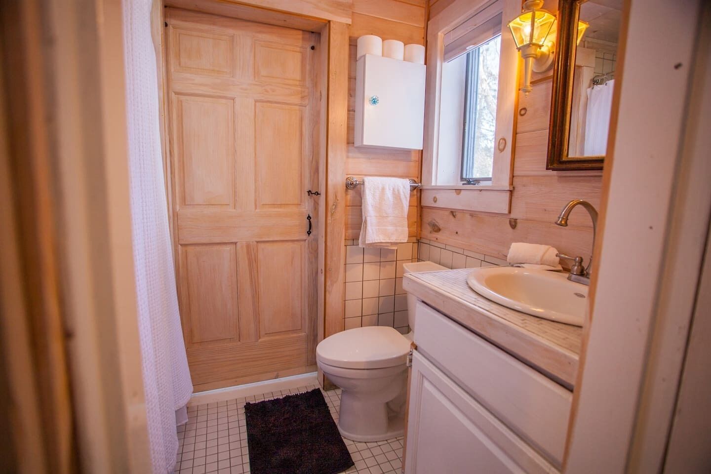 A small bathroom with a toilet, sink, and mirror. There is a towel rack above the sink and a door with a towel hanging on it.
