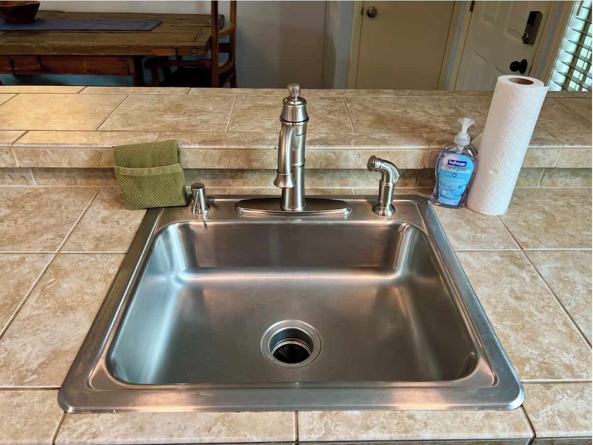 A stainless steel kitchen sink with a green towel and a soap dispenser on the countertop.