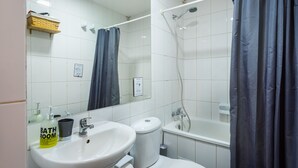 Un baño con lavamanos, inodoro y bañera blancos. Hay una cortina de ducha, un espejo y una repisa con una cesta.