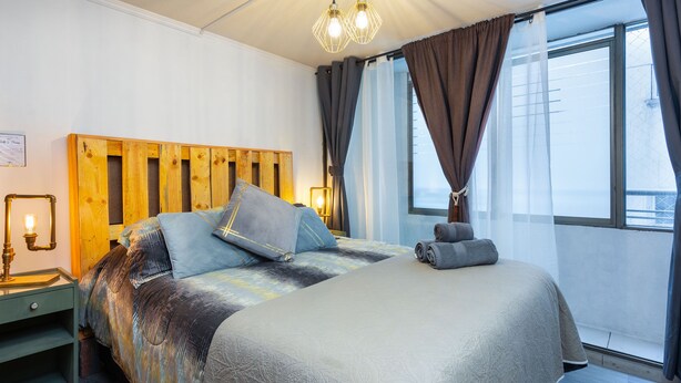 Una cama bien hecha con cabecera de madera, una mesita de noche con lámpara y vistas al exterior a través de un ventanal grande.