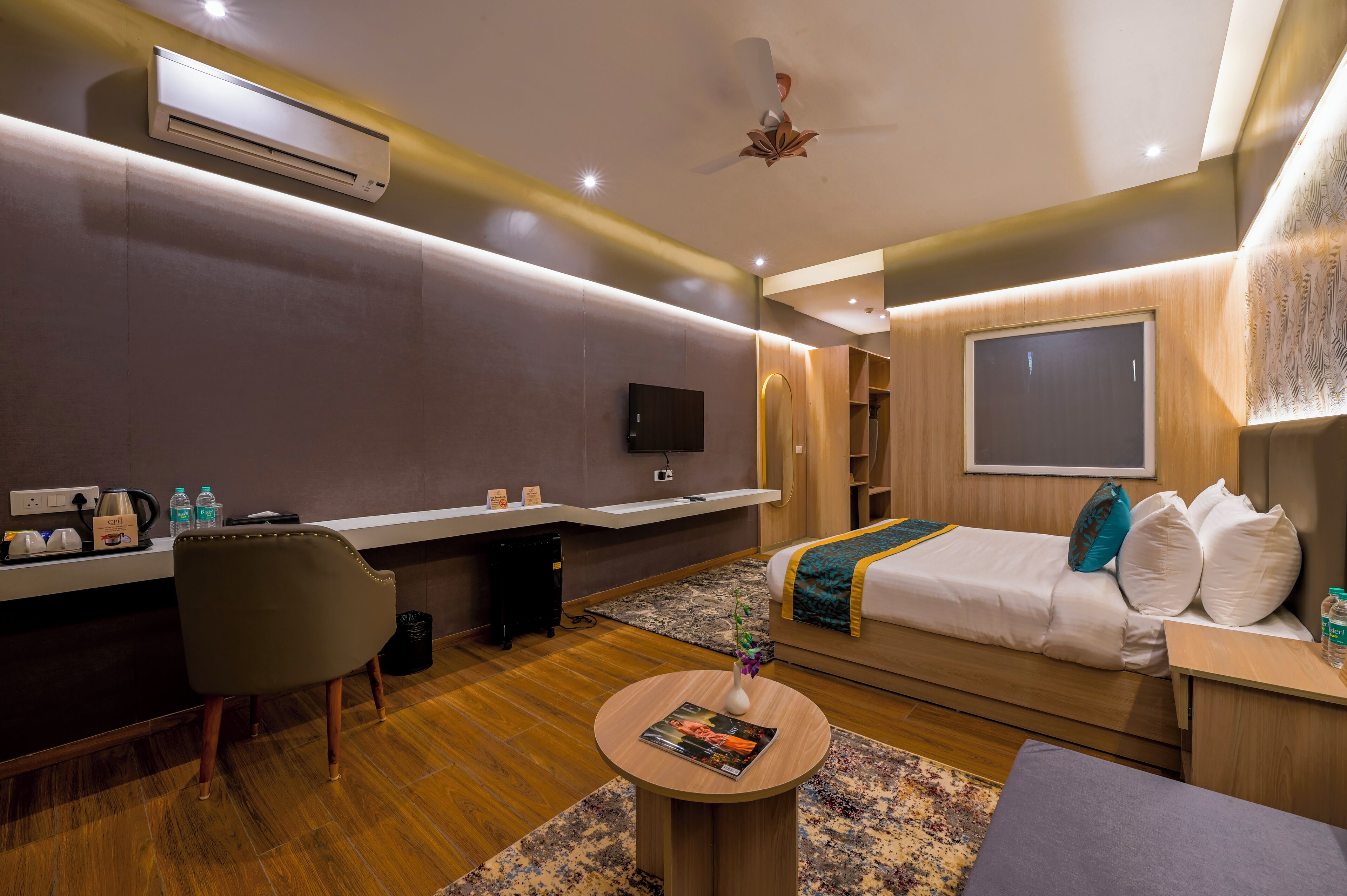 Una habitación de hotel moderna con cama, mesita de noche, silla y una pequeña zona de escritorio con televisión de pantalla plana y cafetera.