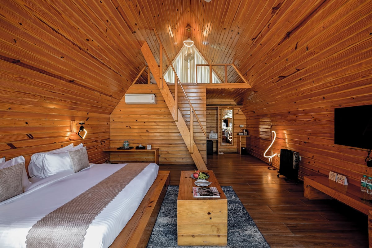 A log cabin interior with a wooden staircase leading to a loft bedroom, a central dining area, and a living space.