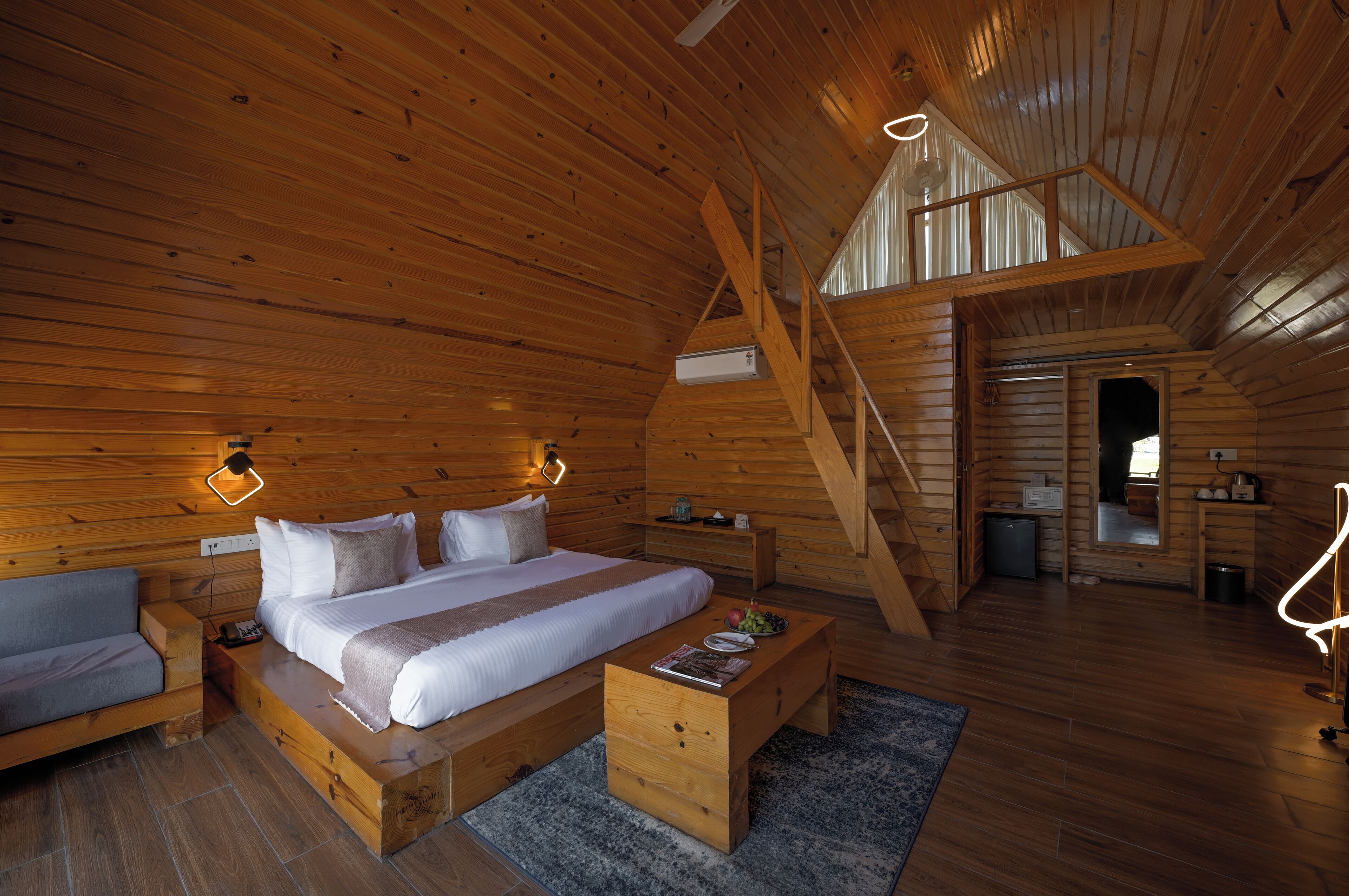 A wooden cabin bedroom with a bed, bedside tables, a sofa, a wooden coffee table, and a staircase leading to an upper level.