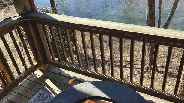 A round outdoor table with a fire pit in the center, surrounded by a wooden deck and a view of a body of water.