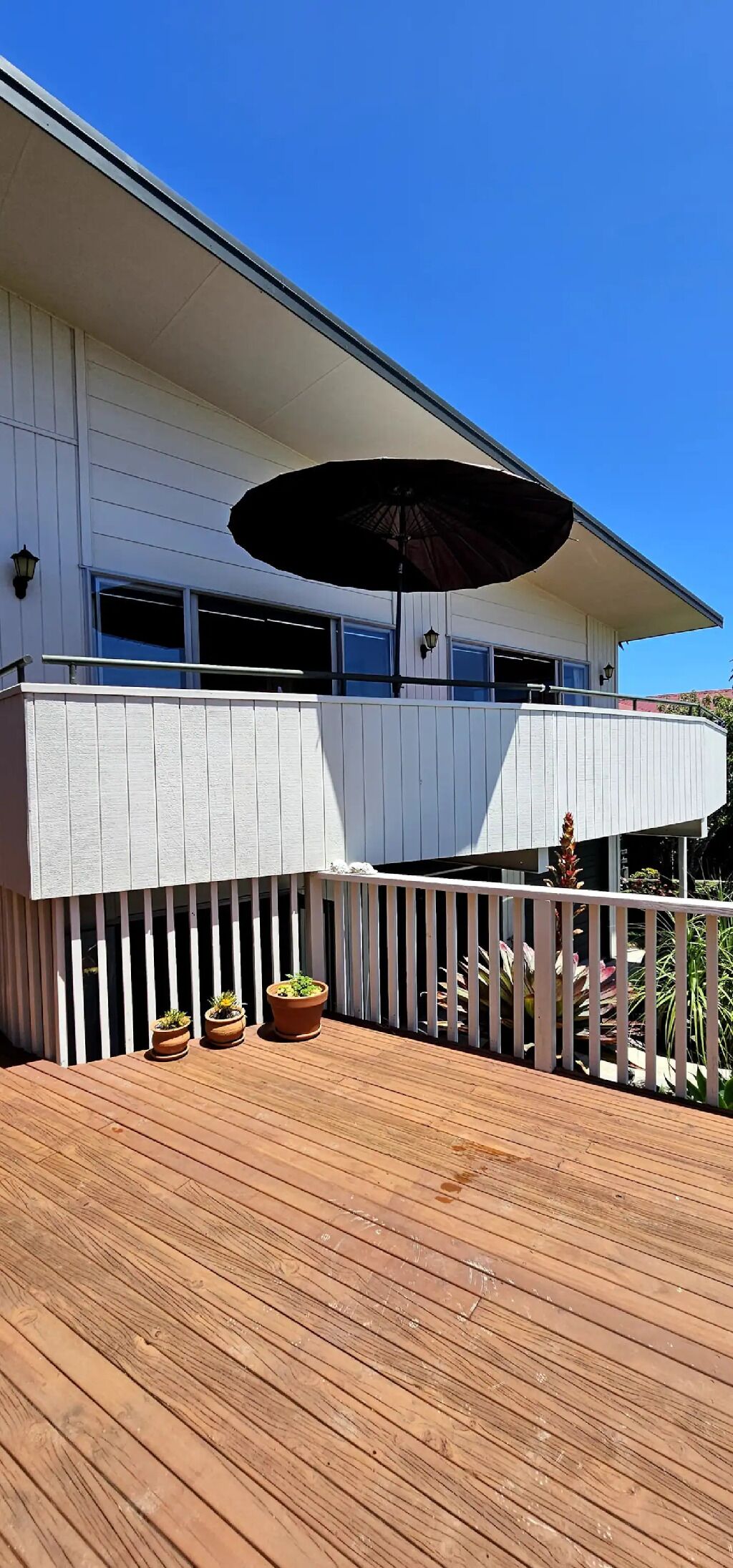 Une terrasse en bois agrémentée de plantes en pot, d’un grand parasol et d’un bâtiment blanc doté d’un balcon.