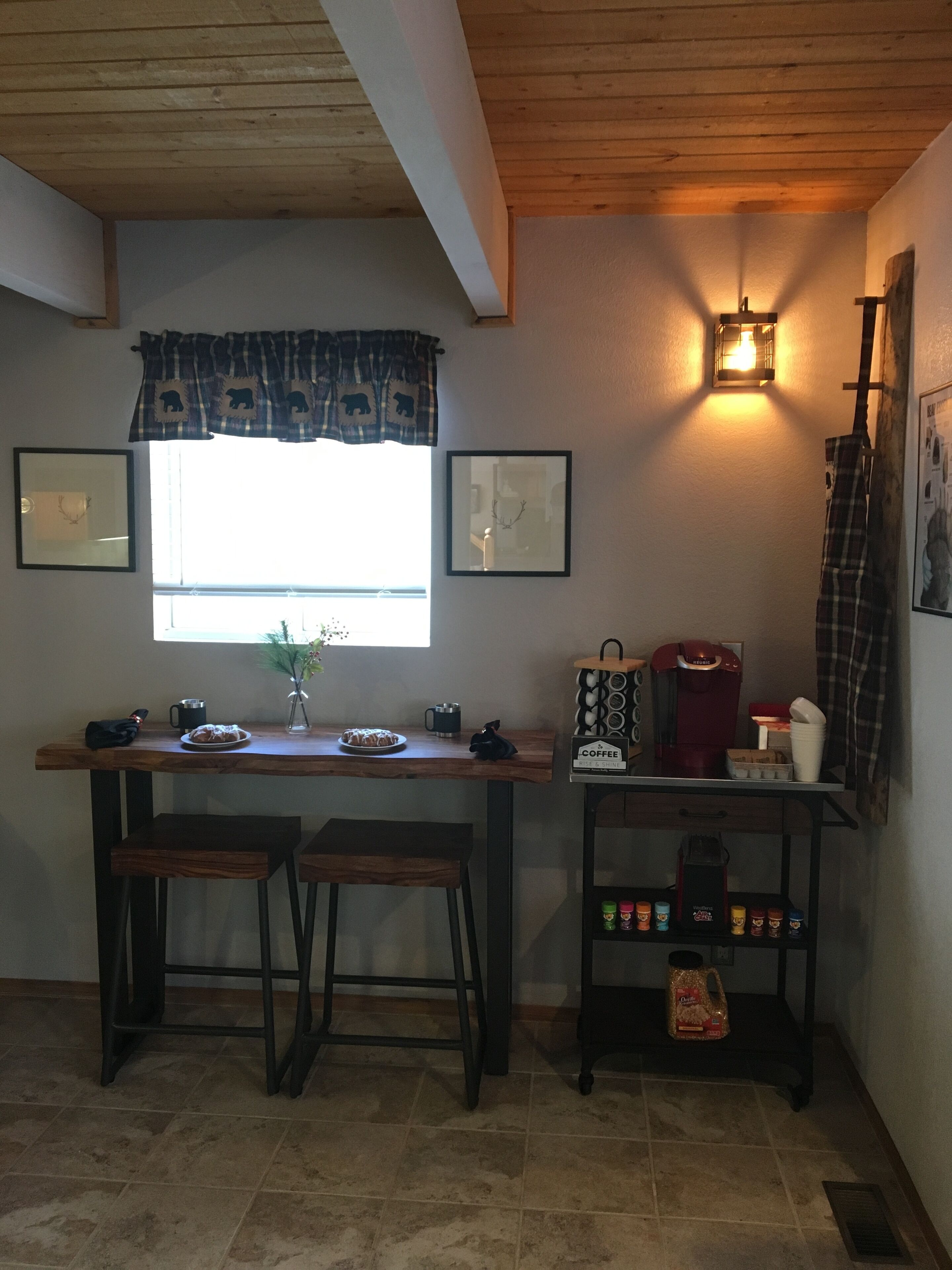 Una cocina acogedora con una mesa de madera, dos taburetes, una cafetera y un estante con diversos objetos.