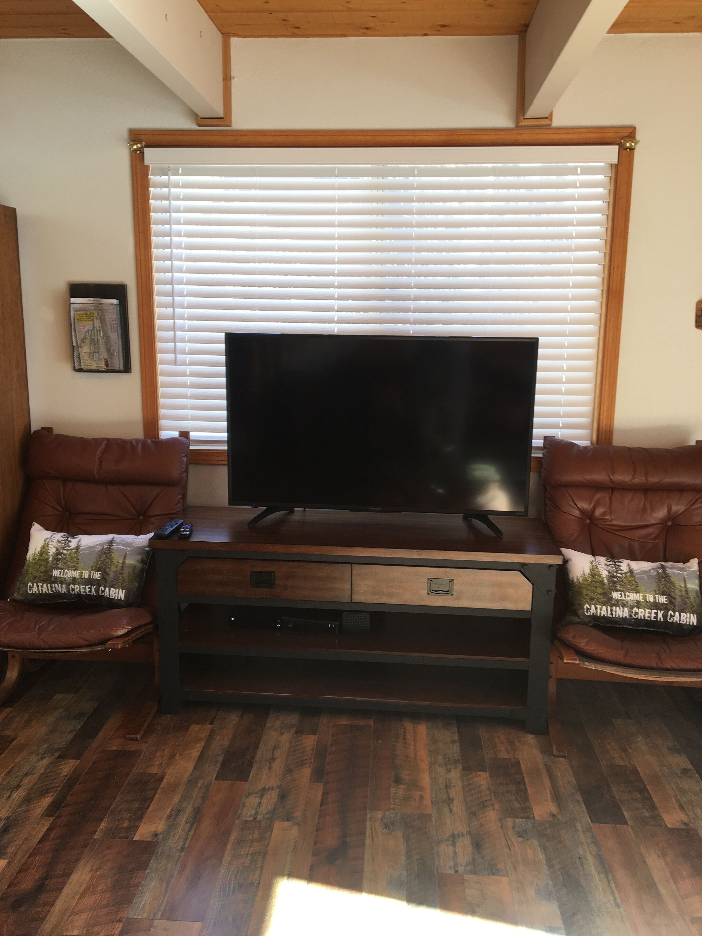 A living room with a flat-screen TV on a wooden stand, two brown leather sofas with matching cushions, and a window with blinds in the background.