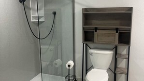 A bathroom with a glass-enclosed shower, a toilet, and a wooden shelf unit. - Rally Rental:  4-bedroom house - walking distance from Sturgis Main Street. (Sturgis)
