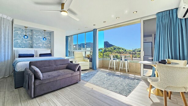 A modern hotel room with a large bed, a grey sofa, a balcony with a table and chairs, and a view of the mountains.