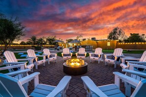 A patio area with Adirondack chairs arranged around a central fire pit, under a sunset sky.