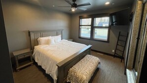 A bedroom with a bed, a bench, a ceiling fan, a window, and a TV mounted on the wall.