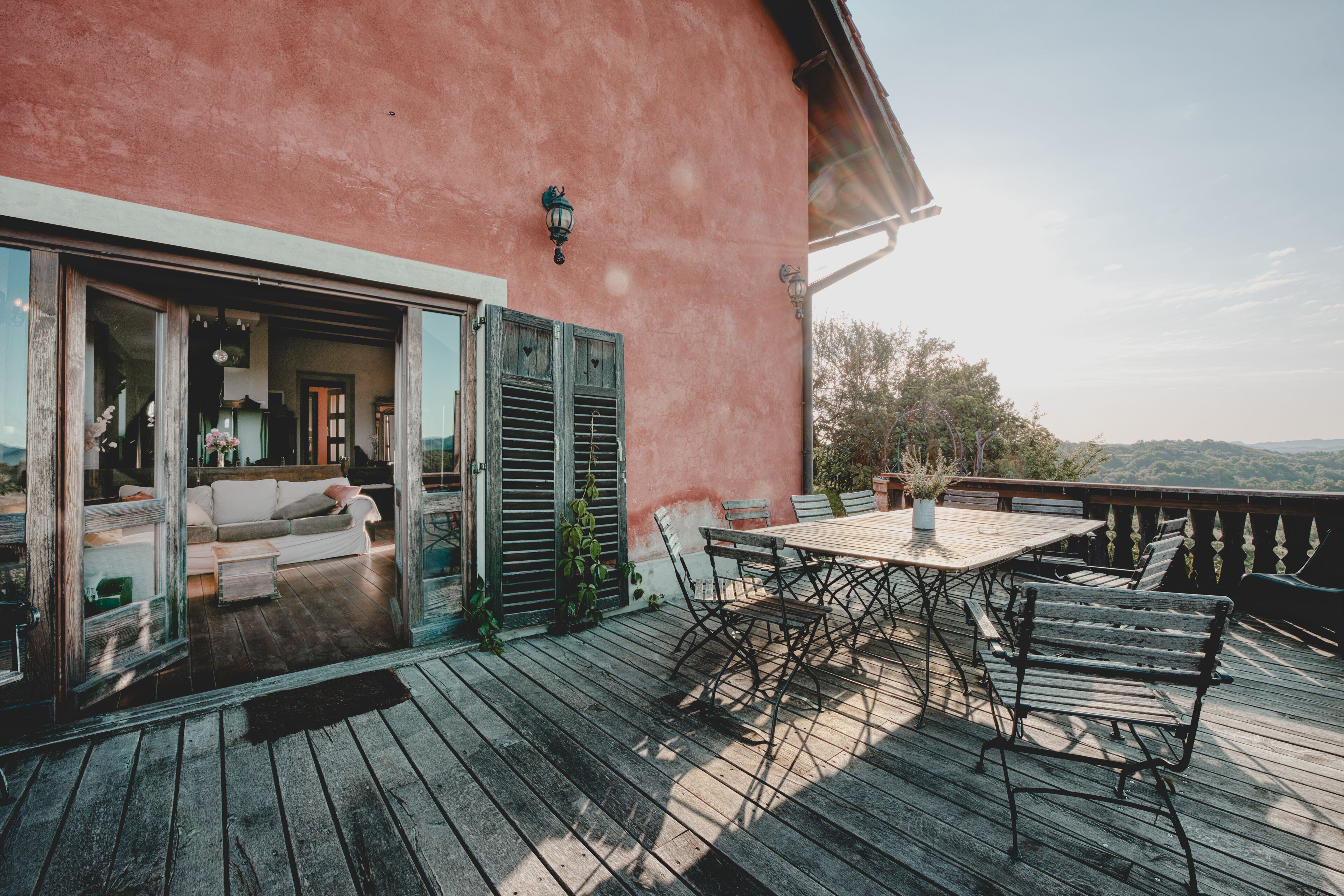 A wooden deck with a table and chairs, a sofa, and a sliding glass door leading to an interior space with a white couch and a potted plant.