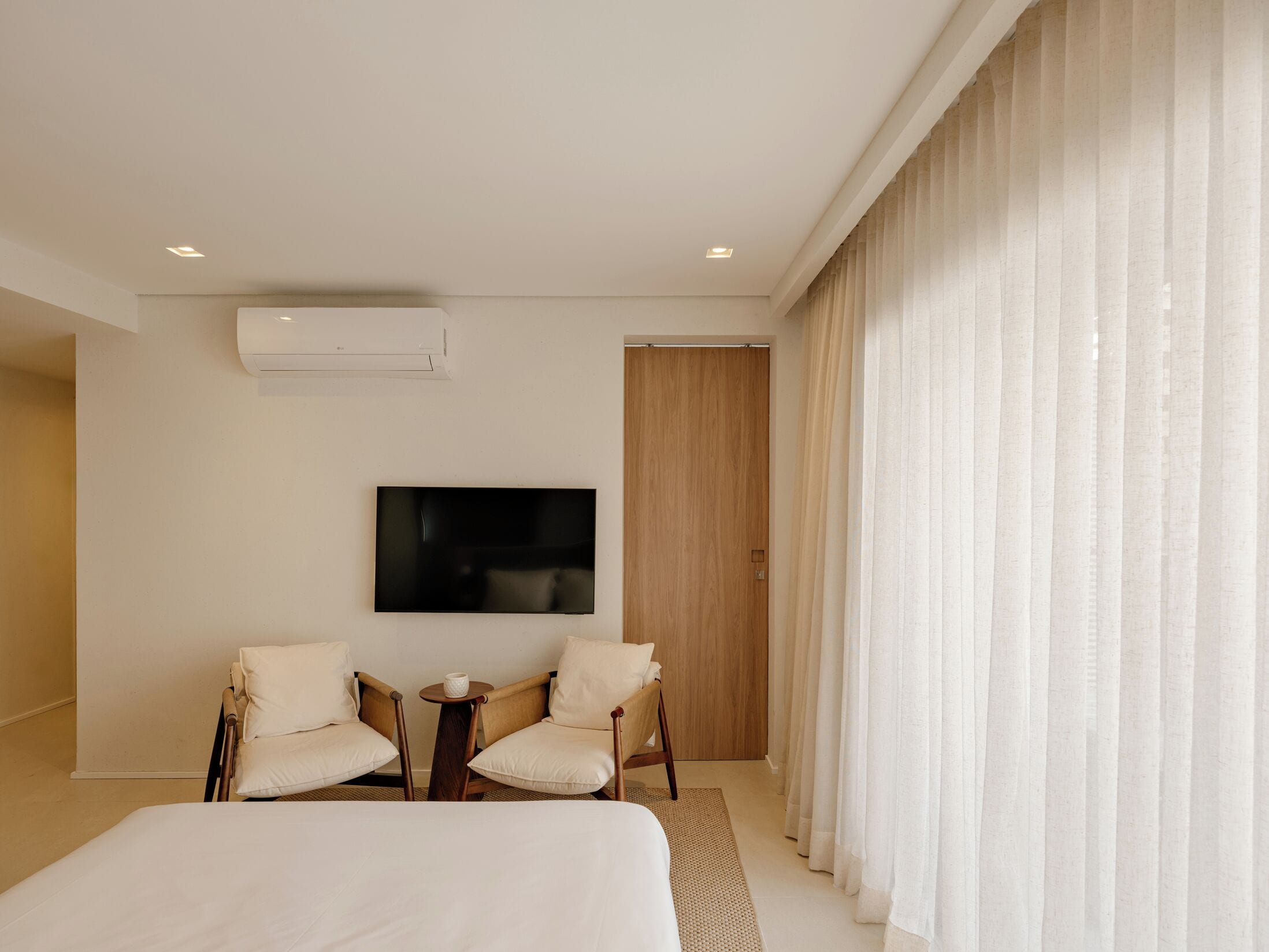 Habitación de hotel moderna con una cama grande, dos sillones, una mesita, un televisor de pantalla plana y un amplio ventanal con cortinas translúcidas.