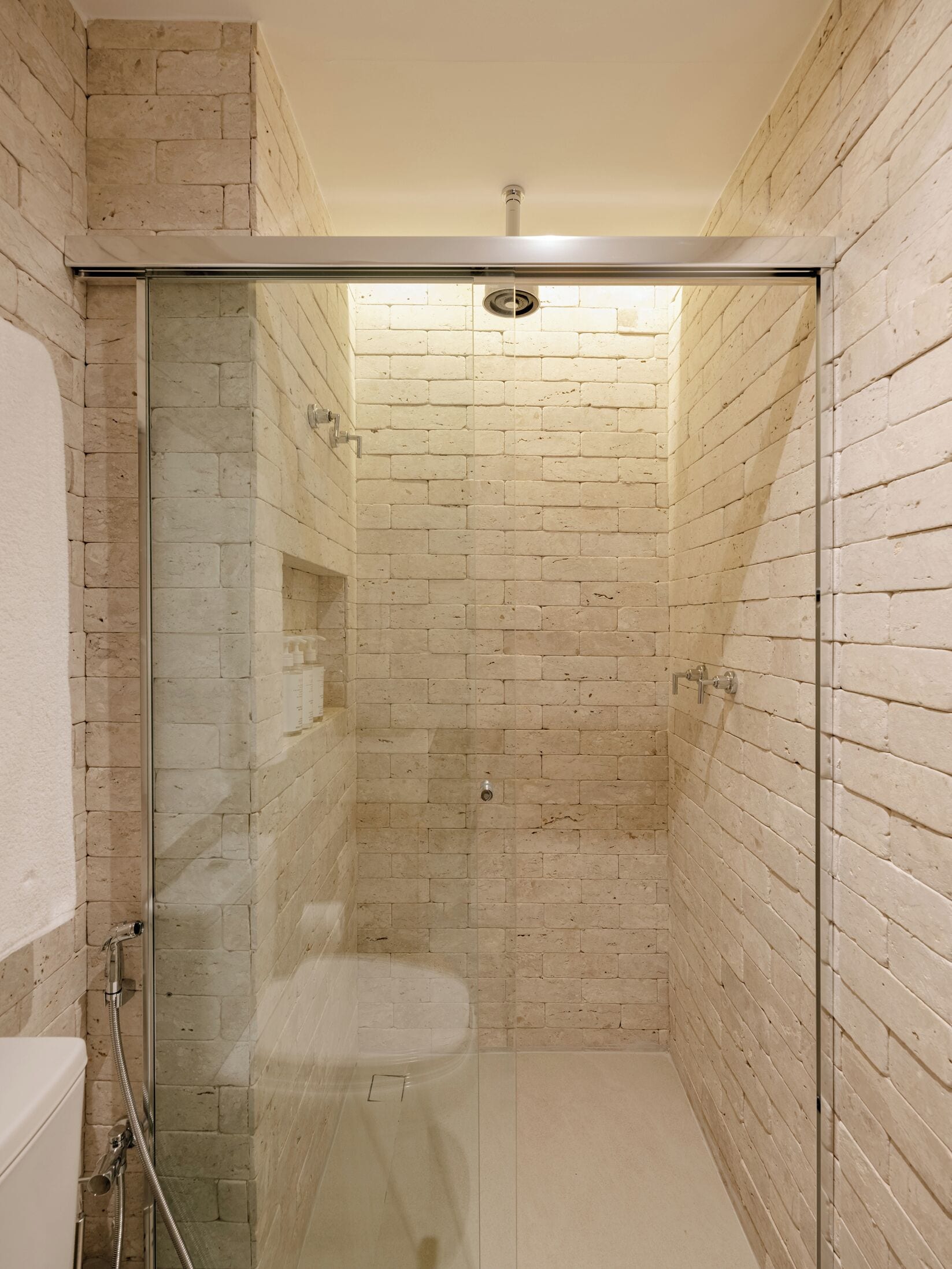 A modern bathroom with a glass-enclosed shower, beige brick walls, and a white ceiling.