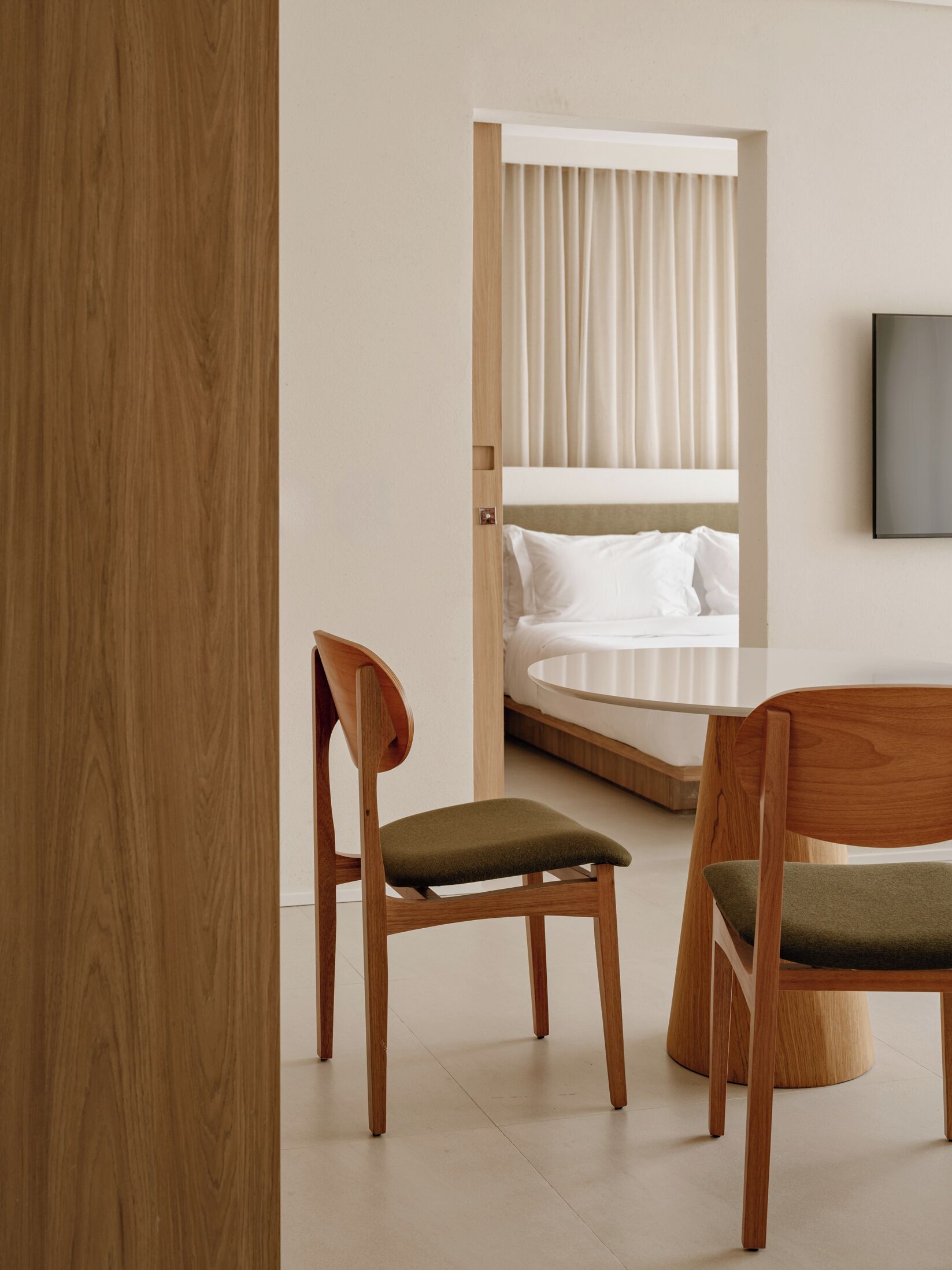 Una habitación de hotel moderna con suelo de madera, una cama con ropa blanca y dos sillas de madera con cojines verdes.